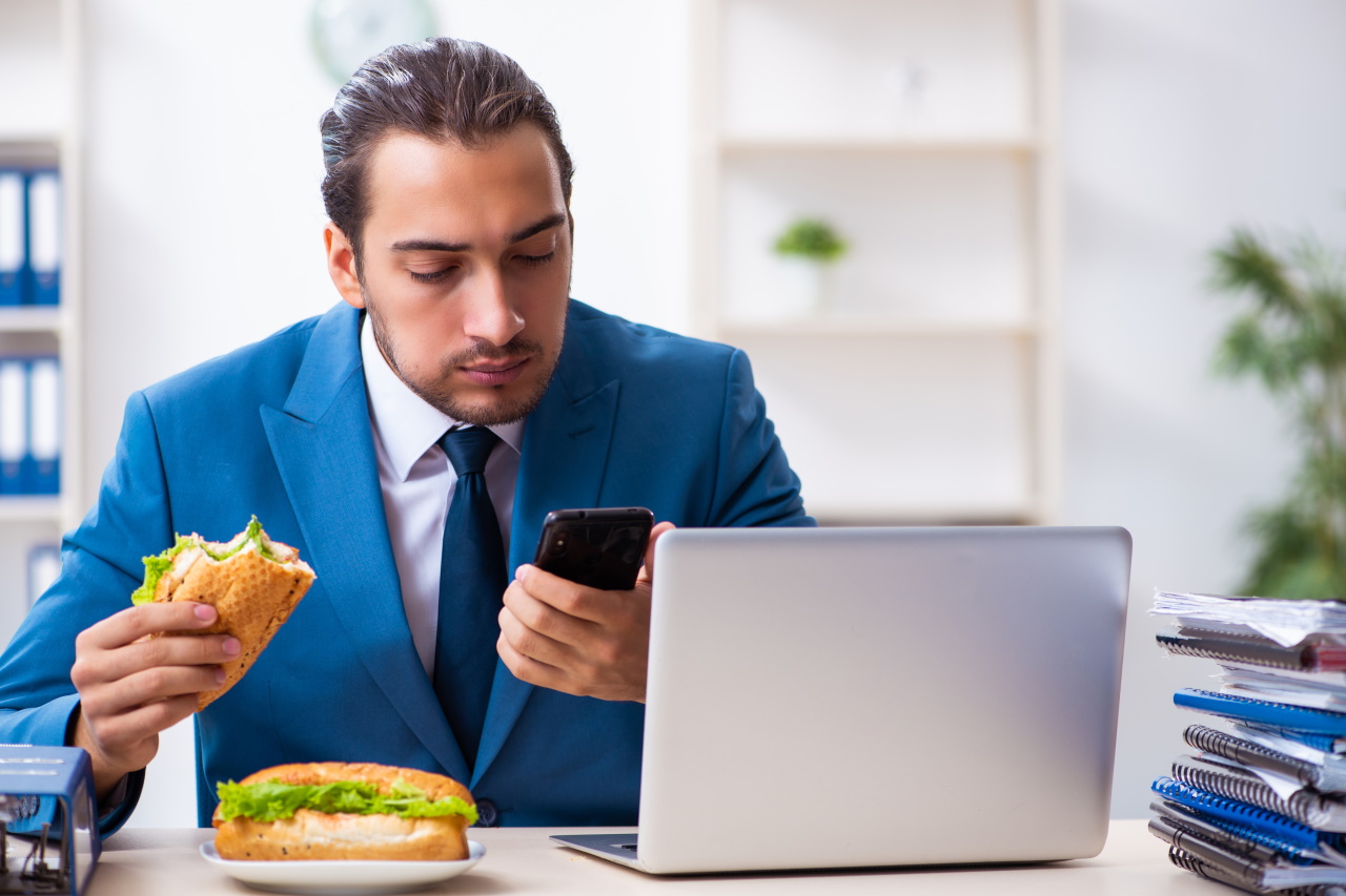 Mężczyzna w garniturze siedzi przy biurku z telefonem w dłoni i otwartym komputerem - je kanapki z sałatą