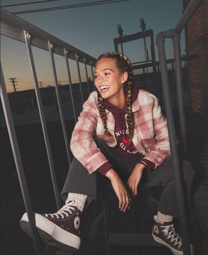 młoda uśmiechnięta dziewczyna z warkoczami siedzi na metalowych schodach w brązowych trampkach Converse