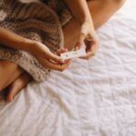 A person is sitting on a bed, holding a pregnancy test showing a positive result.